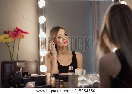 Attractive Blonde Girl Primp At The Mirror With Lamps In The Beauty Studio.