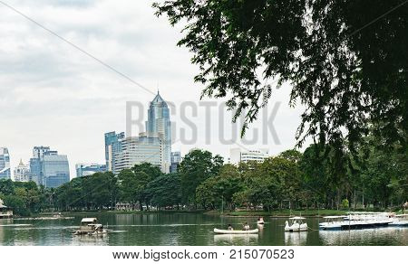 Bangkok, Thailand - December 23, 2015: People relax on boat in a city park. Pleasureboats on a lake in the heart of Bangkok - Lumphini Park. Business district cityscape with cloudy sky in the evening