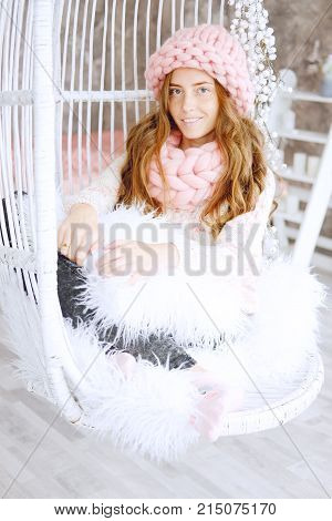 Young woman with red hair and dressed in sweater and knitted pink hat and snood sitting in her favourite chair