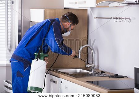 Pest Control Worker In Uniform Spraying Pesticide With Sprayer In Kitchen