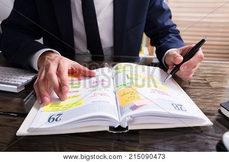 Close-up Of A Businessperson's Hand Writing Schedule In Diary At Workplace