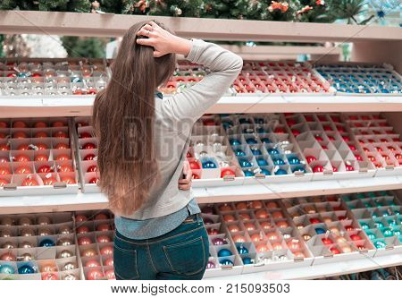 Cheerful happy customer girl choosing gifts for Christmas and New Year. She is taking red Christmas tree decoration balls. difficult decision. Various choice