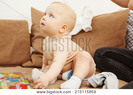 Profile of innocent infant with perfect skin stands on arms and legs on sofa with broad smile. Adorable newborn child in pants