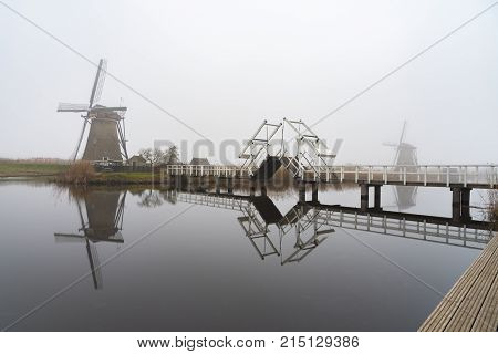 Silhouette Of A Dutch Windmill Under A Misty And Foggy Weather A Long A Calm And Quiet Canal