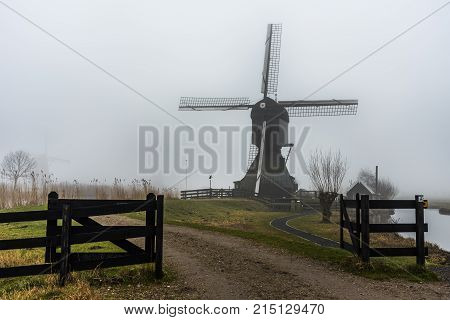 Silhouette Of A Dutch Windmill Under A Misty And Foggy Weather A Long A Calm And Quiet Canal