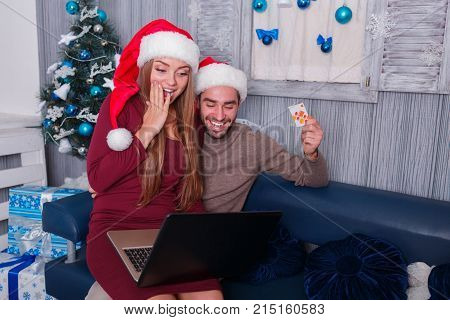 Happy young couple smiling, doing online shopping, on New Year's Eve, sitting on sofa, using credit card and laptop, the little one is sitting on his knees with a man covering his mouth with his hand.