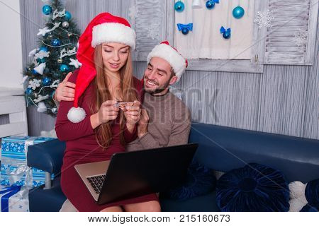 Happy young couple smiling, doing online shopping, on New Year's Eve, sitting on sofa, using credit card and laptop, the little girl is sitting on the lap of a man looking at a credit card.