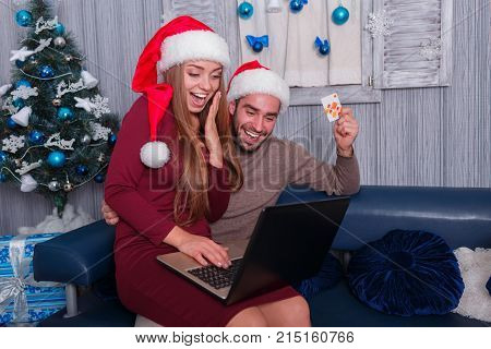 Happy young couple smiling, doing online shopping, on New Year's Eve, sitting on sofa at home, using credit card and laptop, the little one is sitting on his lap beside the man.
