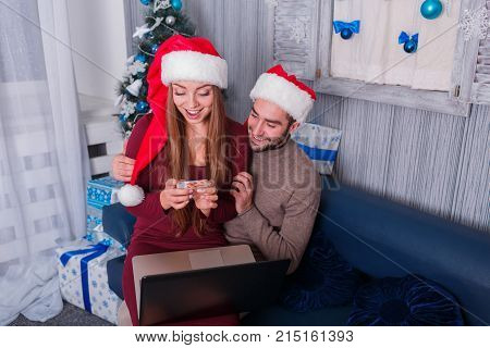 Happy young couple smiling, doing online shopping, on New Year's Eve, sitting on sofa, using credit card and laptop, the little girl is sitting on the lap of a man looking at a credit card.