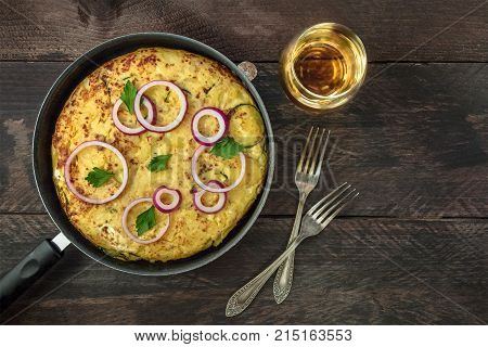 An overhead photo of a Spanish tortilla with potatoes and zucchinis in a tortillera, with a glass of white wine, two forks, and a place for text