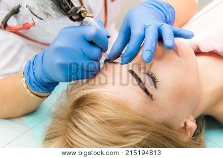 Cosmetologist applying permanent make up on eyebrows. Beautiful woman with smooth young skin sitting in a beautician. A beautician performs modeling permanent eyebrows makings.