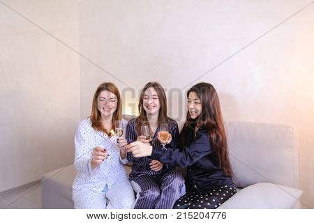 Three wonderful women having fun and drinking white wine or champagne from glass goblets, laughing and talking on girlish themes, discussing guys sitting on gray soft sofa in bright bedroom at night. 
