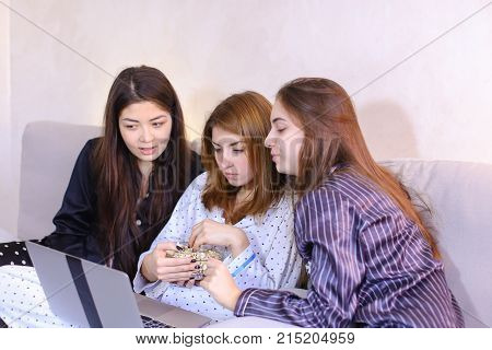 Three wonderful women have fun, look at computer comedy and eat popcorn, laugh and talk on girlish themes, pose in camera with smile on their face, sitting on gray soft sofa in bright bedroom at night. European-looking girl with medium-length fair-haired 