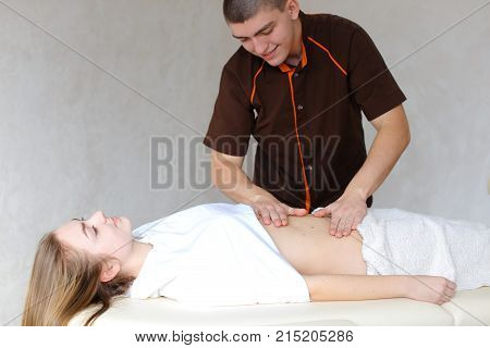 Friendly and professional guy doctor and masseur conducts strengthening and healing procedure for abdomen and body as whole, communicates and tells useful medical information that came to girl who lies on couch up to half covered with terry towel in light