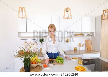 Stunning young woman slices fresh green cucumber for dinner with table knife, picks up and senses freshness of lettuce leaves and continues shredding vegetables. Blonde at top of modern light kitchen with bowl of fruit and vase with potted plant and dress