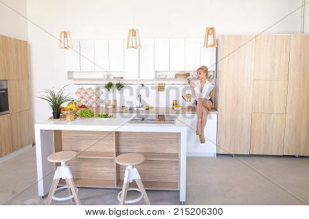 Marvelous woman holds mobile in hands and reads articles on cooking or scrolls news on social networks, pulls out of plate and tastes potato chips sitting on kitchen surface of stylish white kitchen with oven and variety of cooking accessories. 