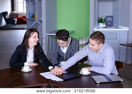 Cheerful and active staff of company, two men and women, with smiles on their faces and in good spirits, completed work process and lay their hands on table, folding each other, palms down and raising them upwards, screaming and raising team spirit, sitti