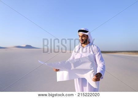 Beautiful Emirate Sheikh businessman studying project of future construction of complex, standing in middle of bottomless desert with white clean sand on clear warm day against blue sky. Swarthy Muslim with short dark hair dressed in kandura, long, spacio