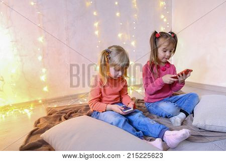 Lovely little girls holding mobile phones and playing games or taking pictures,  typing text for messages on touch screen, they laughing and smiling, sitting on rug in bright room with flickering New Year's garland in evening. Blond girl of European appea