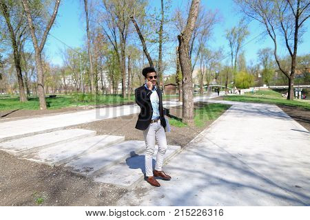 Guy walking in park and smiling, slow motion. Man dressed in blue shirt, white jeans and brown shoes. Boy has black hair and beard. Concept of places for rest and clean parks and cheeps fashion clothes for man