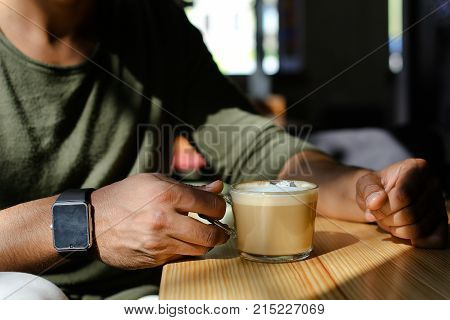 Close-up shooting, man sitting in cafe and drinking hot coffee with milk. Boy dressed in khaki pullover. Attractive mullato has dimples, beard, full lips and black hair. Concept of modern places for young people cheap prices.