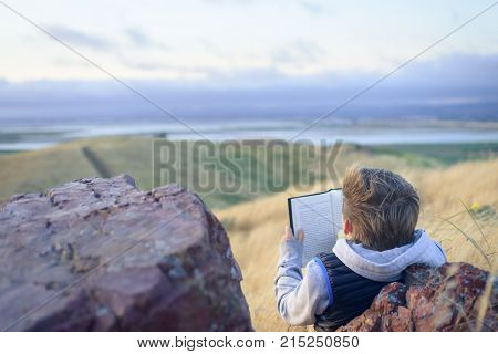 Boy reading book outside on the top of the hill, up above the ocean, unschooling education