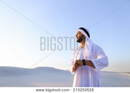 Attractive guy, emirate businessman uses for business and for work iWatch, who are put on hand, smiling and examining landscapes of large sandy desert against blue sky on hot summer morning.