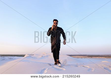 Modern Muslim chats on smartphone with smile on face and bounces on spot, spreading arms and legs to sides, being in bottomless desert with white and clean sand on clear spring evening. 