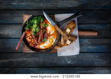 Breakfast in on a hot frying pan with fried eggs, sausages, beans, greenery and toasts. On a colored wooden background with cutlery. Top view.