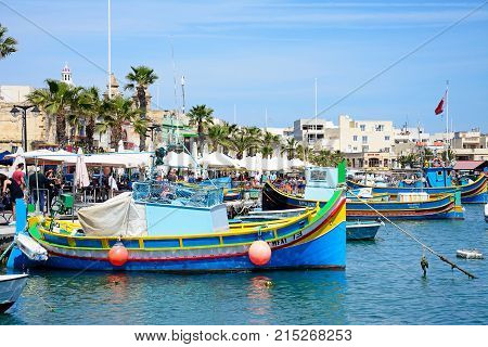 MARSAXLOKK, MALTA - APRIL 1, 2017 - Traditional Maltese Dghajsa fishing boats in the harbour with waterfront buildings and market stalls to the rear Marsaxlokk Malta Europe, April 1, 2017.