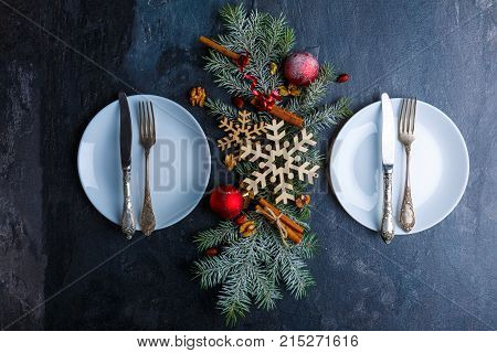On the dark surface, two plates with cutlery and between them Christmas tree is decorated with wooden snowflakes, Christmas balls and cinnamon sticks. View from above. Indoors.