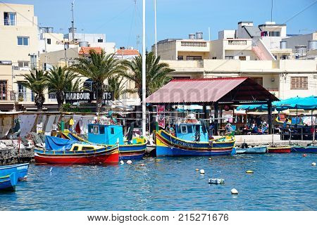 MARSAXLOKK, MALTA - APRIL 1, 2017 - Traditional Maltese Dghajsa fishing boats in the harbour with waterfront buildings and market stalls to the rear Marsaxlokk Malta Europe, April 1, 2017.