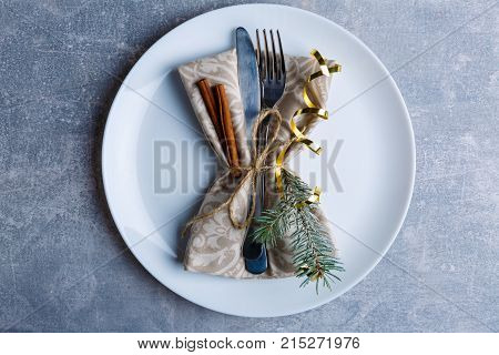 On a round white plate, served cutlery is decorated with a Christmas tree branch, cinnamon sticks and a golden ribbon. Christmas atmosphere. View from above. Indoors.