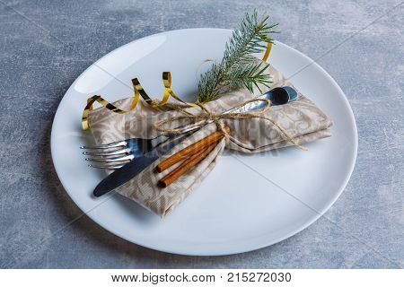 A decorated Christmas table with a round white plate with cutlery wrapped in a tissue napkin decorated with a Christmas tree branch, cinnamon sticks and a golden ribbon. Christmas atmosphere. View from above. Indoors.