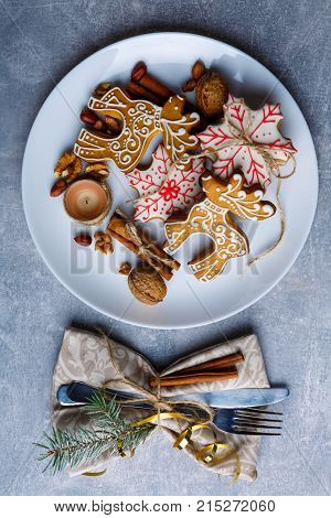Delicious Christmas cookies of different shapes with sugar icing on a white round plate, next to eat cutlery wrapped in a tissue napkin decorated with a Christmas tree branch, cinnamon sticks and golden ribbon. Christmas atmosphere. Indoors.