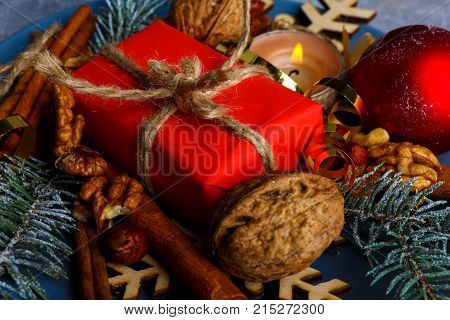 A small gift box in red packing next to a burning candle, walnuts and cinnamon sticks. Christmas decor. View from above. Indoors.