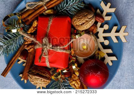 On a blue plate there is a small red gift box, a burning candle, walnuts and cinnamon sticks. Close-up. Christmas decor. View from above. Indoors.