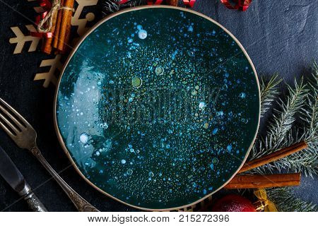 Creative plate in green glossy surface, next to cutlery, decorated with a Christmas tree branch and cinnamon sticks. Close-up. Indoors.