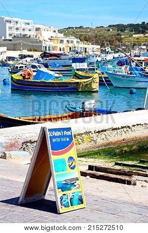 MARSAXLOKK, MALTA - APRIL 1, 2017 - Traditional Maltese Dghajsa fishing boats in the harbour with waterfront buildings to the rear and a water taxi sign in the foreground Marsaxlokk Malta Europe, April 1, 2017.