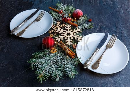On the table decorated with snowflakes and balls Christmas tree branch between two white and round plates with cutlery.