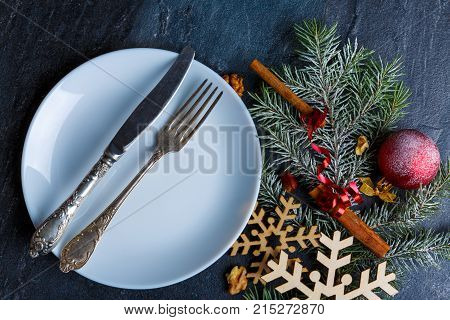 On a white round plate an old knife and fork, near Christmas decorations from a Christmas tree branch is decorated with wooden snowflakes, red balls and fragrant cinnamon sticks.