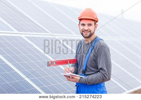 A smiling employee of a solar cell station in uniform and helmet stands with a waterpas in his hands and writes something into a notepad.