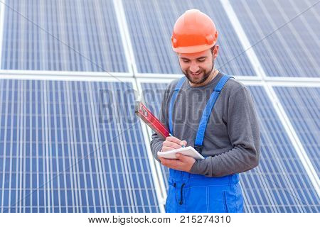 A bearded solar station worker in blue uniform and in an orange helmet, with a smile, writes something in a notebook holding waterpas in his hands.