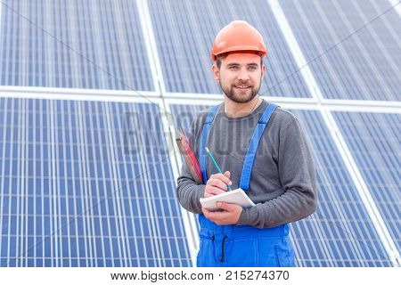 A young worker at a solar power station in blue uniform and in an orange helmet looks somewhere and writes something into a notebook holding waterpas in his hands.