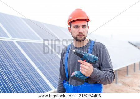 A serious employee of the solar cell station in a gray sweater, blue uniform and in an orange helmet, holds a drill in his hands.