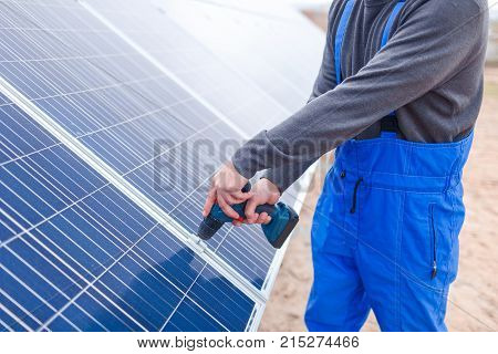 A solar station worker in blue uniform drills a solar battery panel. Outdoors.