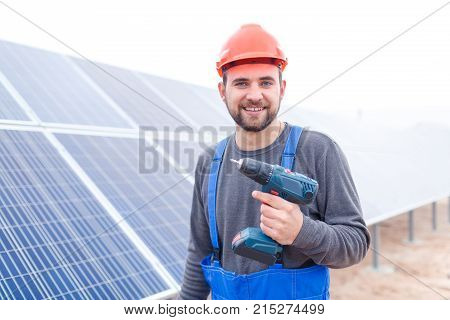 A man with a beard worker at a solar station in a blue uniform is smiling and holding a drill. Outdoors.
