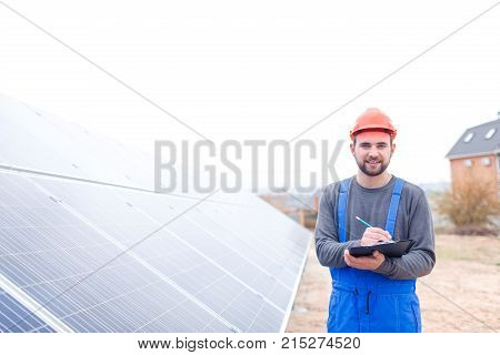 A solar station worker in blue uniform and an orange helmet stands near the solar battery panel smiling and writes something into a notepad. Outdoors.