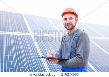 A solar station worker in uniform and a helmet stands on the background of a large panel of solar battery and records something in a notebook and smiles. Outdoors.
