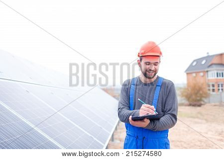 A happy worker stands at the solar station in uniform and helmet and writes something in a notebook with a smile. Outdoors.